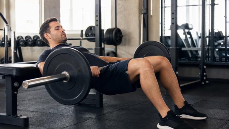 Man in a black t-shirt and shorts performing a barbell hip thrust exercise with his back against a weight bench in a bright gym.